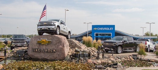 A silver Chevrolet Silverado pickup truck prominently displayed on a large rock with a 'LIKE A ROCK' inscription outside a dealership.