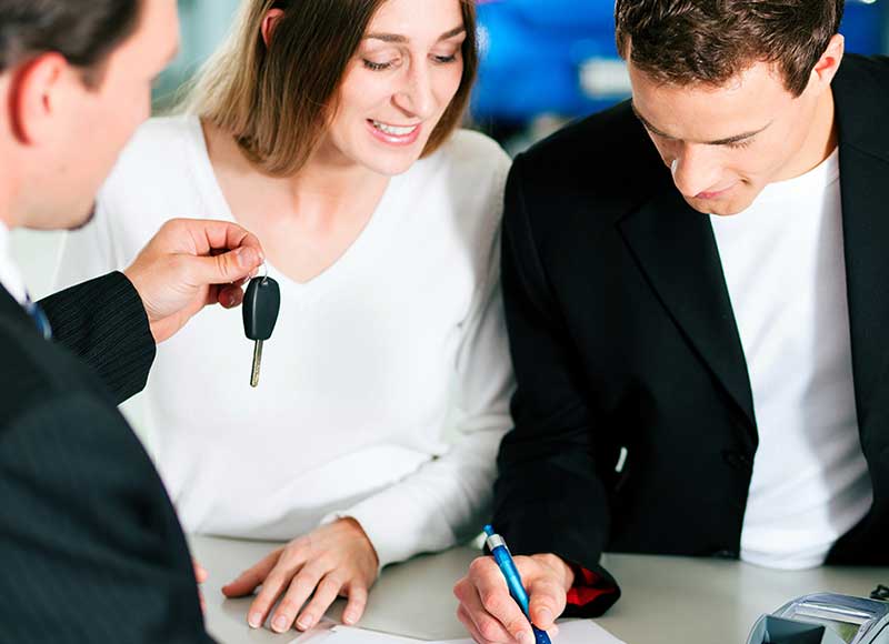 A salesperson hands over car keys to a smiling woman as a man signs a document at a dealership.